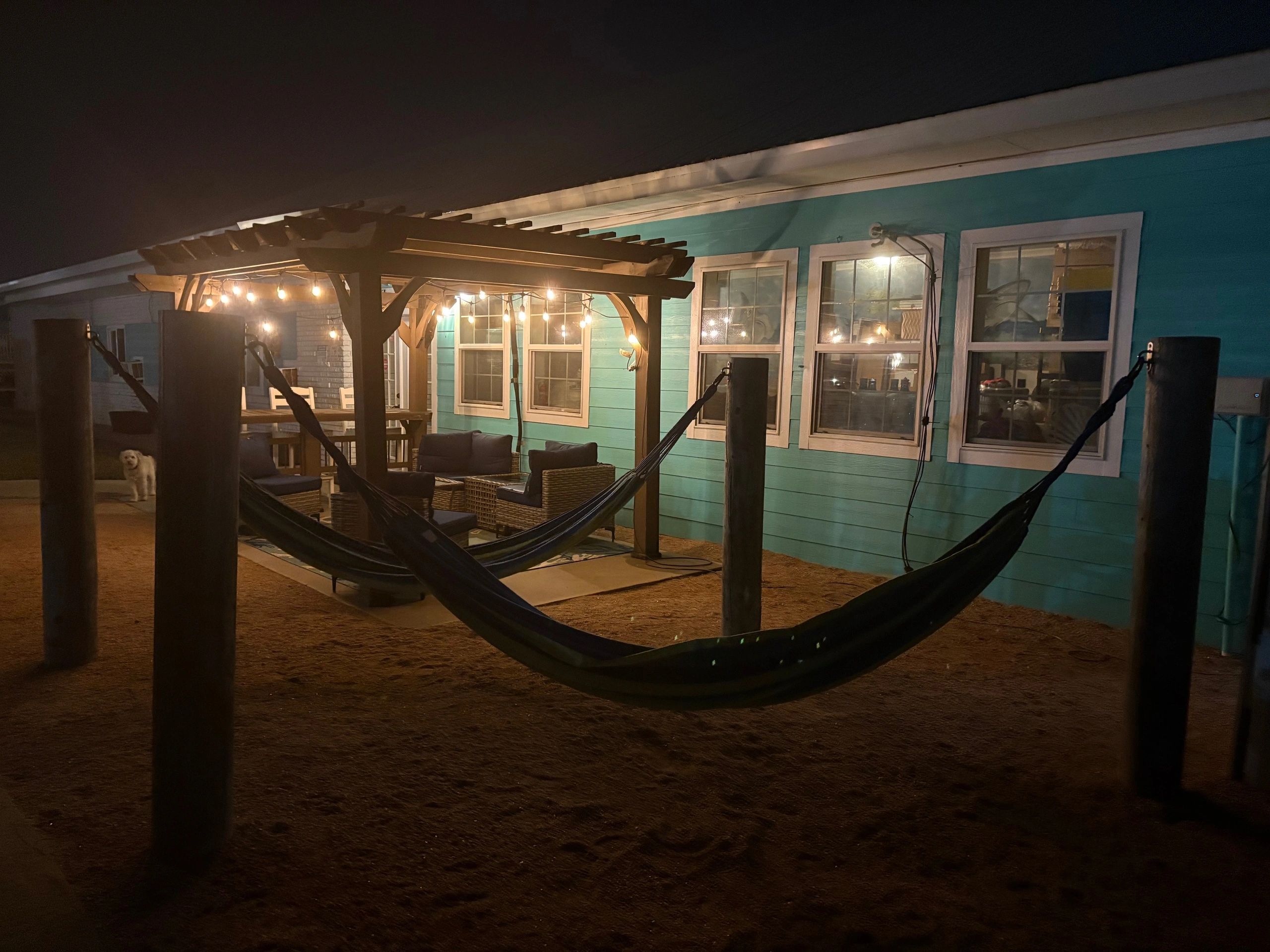 A nighttime view of an outdoor seating area with hammocks, pergola, and string lights in front of a teal building.