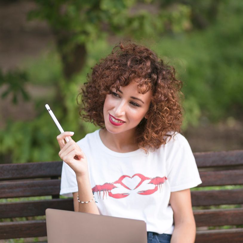 A trendy woman wearing a white t-shirt, sitting on a park bench with her laptop, holding a pen.
