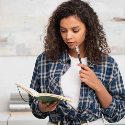 A woman with curly hair journaling, holding a pen and looking pensive.