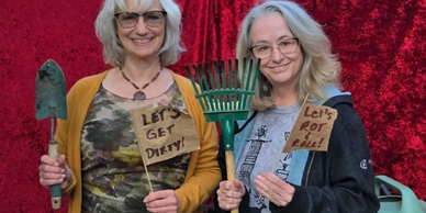Two women holding garden tools and signs with playful gardening slogans.