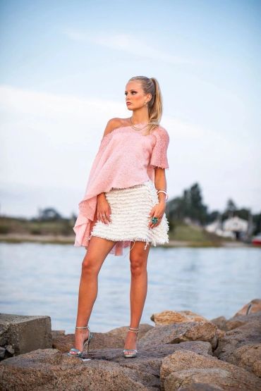 Woman in pink sweater and white skirt posing by the water.