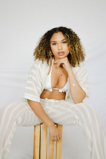 Woman sitting confidently on wooden stool in white striped outfit.