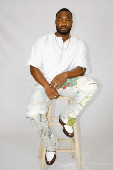 Man sitting on a stool wearing a white shirt and patterned pants.
