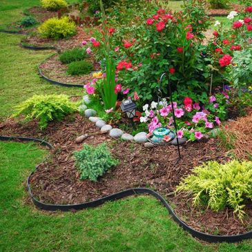 A colorful garden bed with red roses, pink flowers, and green shrubs bordered by black edging.