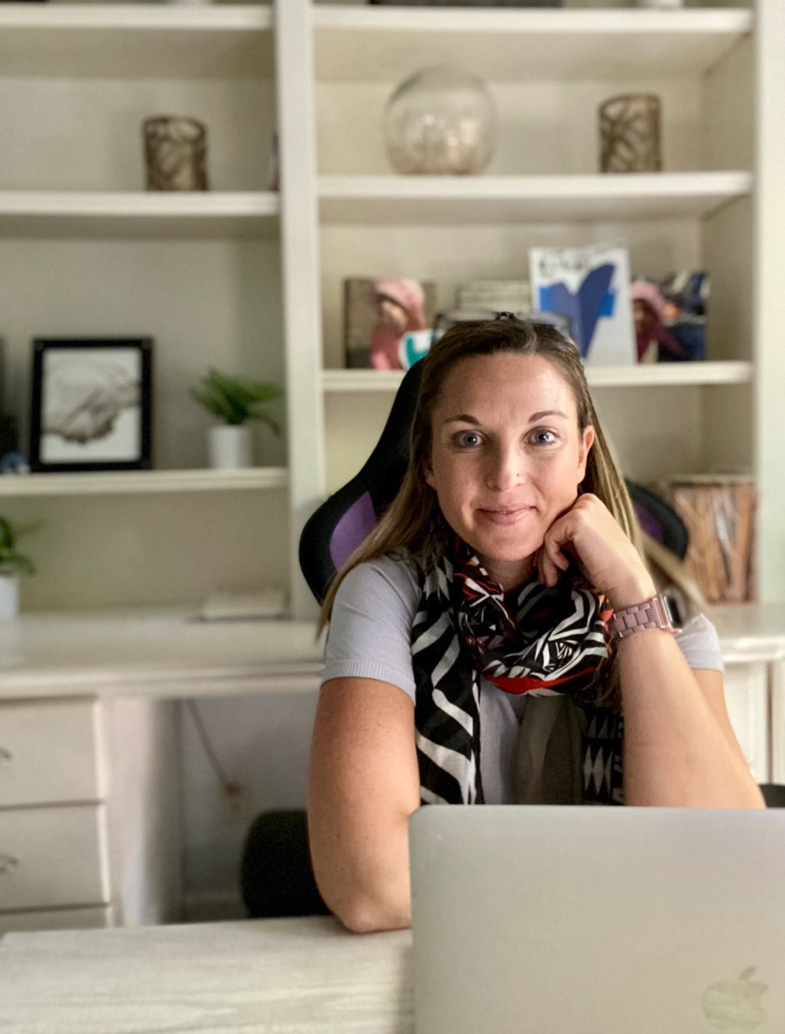 Psychologist sitting at desk where she preforms virtual therapy. Office decor on builtin shelves