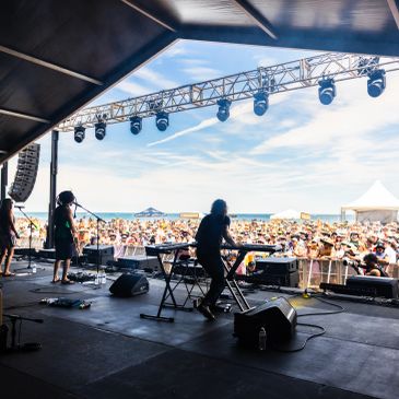 Band performing on stage at an outdoor beach festival with a large audience.