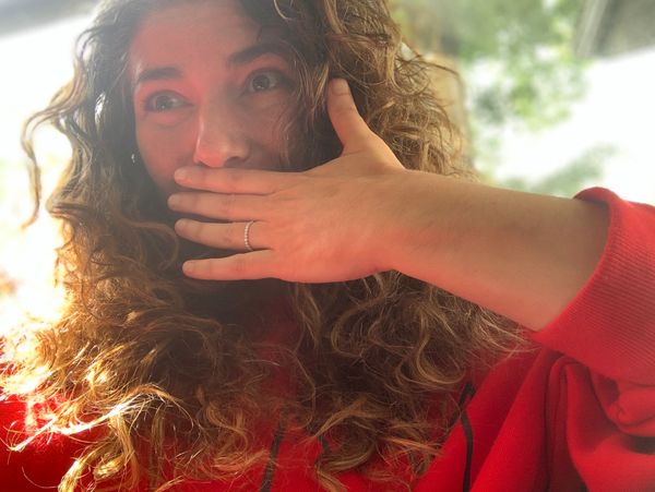 Woman with curly hair covering her mouth, showing a ring on her finger.