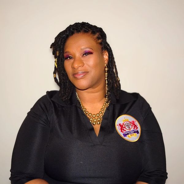 Confident woman with braided hair and gold accessories wearing a black top with an emblem.