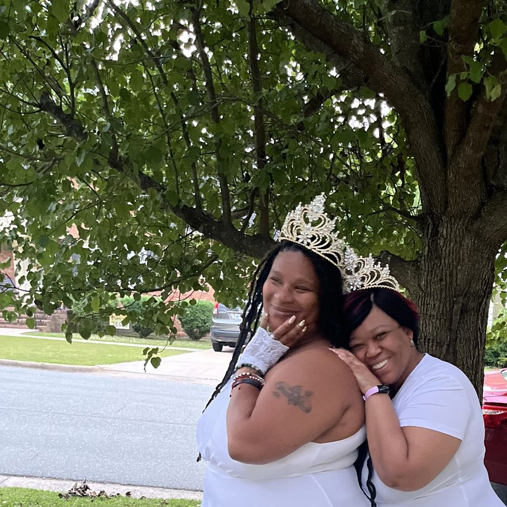 Two women wearing white dresses and ornate crowns smile under a large tree.