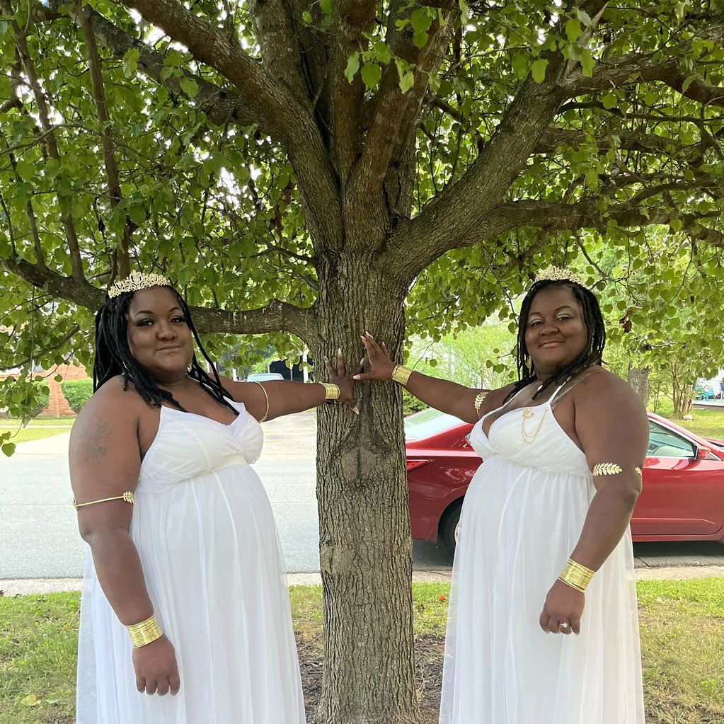 Two women in white dresses and gold accessories pose by a tree outdoors.