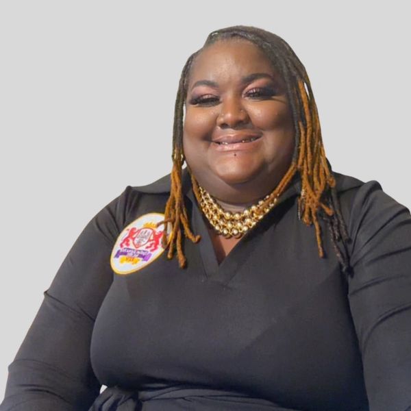 Smiling woman with dreadlocks and gold necklace wearing a black shirt with a patch.