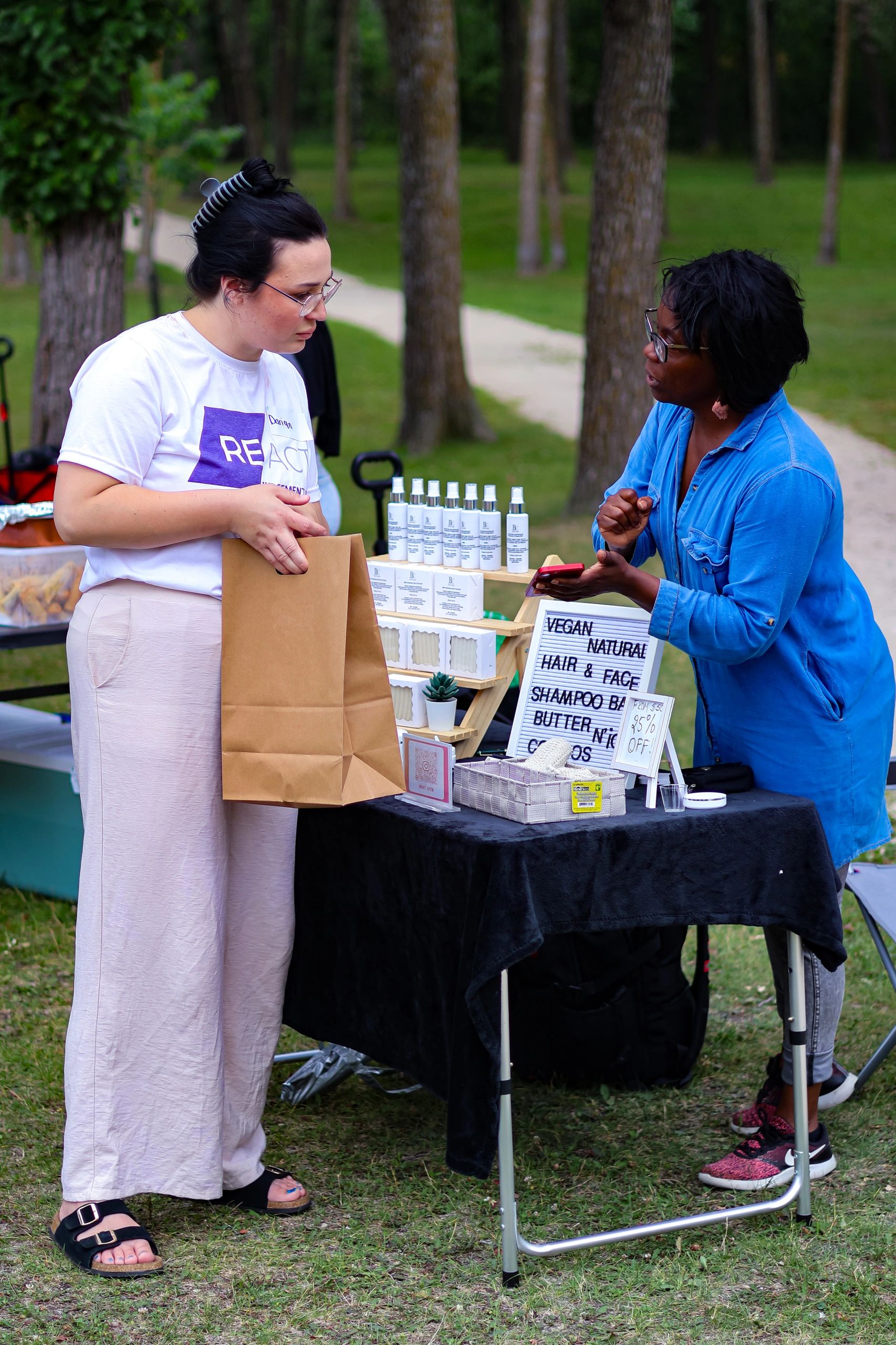 Two women discussing vegan natural hair and face products at an outdoor market stall.