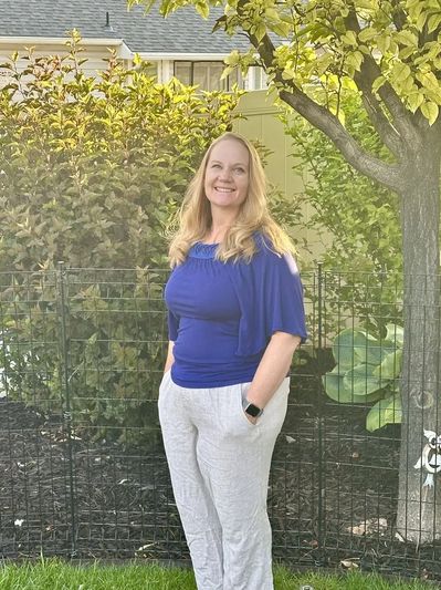 Smiling woman in blue top and light pants stands outdoors by tree and fence.