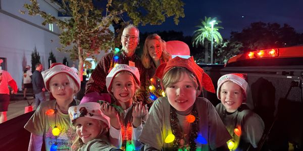Children and adults wearing Christmas hats and colorful lights at night.