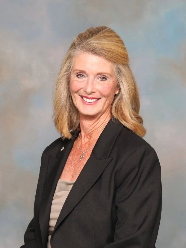 Smiling woman in a black blazer with blonde hair and a cross necklace.