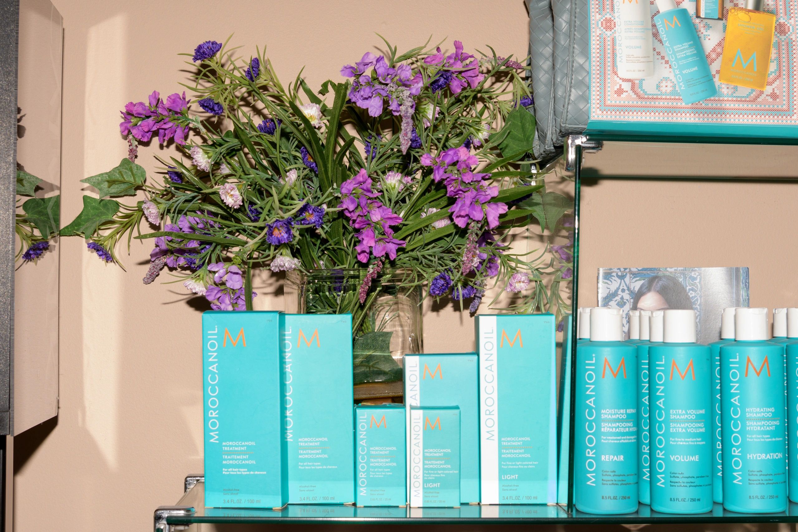 Purple flowers in a vase surrounded by Moroccanoil hair products on shelves.