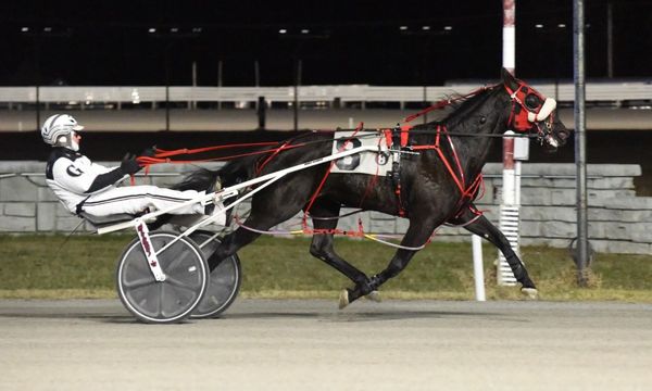 A harness race horse with a driver in white racing gear at night.
