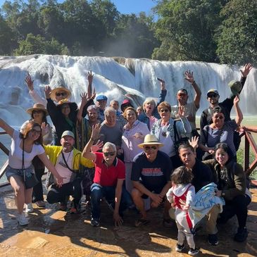 Group of people joyfully posing in front of a waterfall with raised hands.