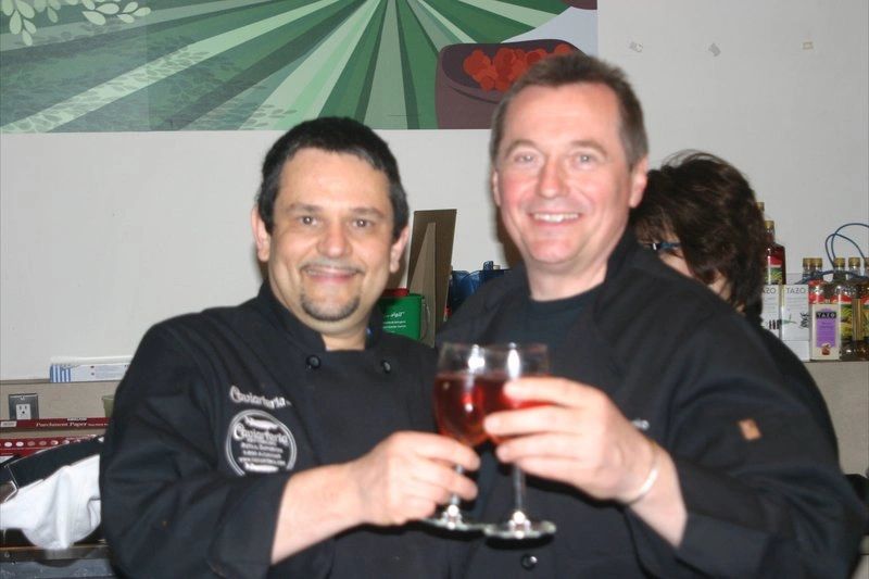 Two chefs clinking glasses with red wine in a kitchen setting.