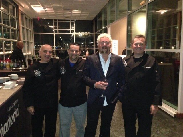 Four men posing inside a well-lit modern venue, three in chef uniforms and one in a suit holding a glass.