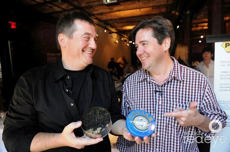 Two men smiling, holding a container of caviar in an indoor setting.
