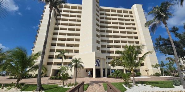 A tall beige building with balconies, palm trees, and a clear blue sky.