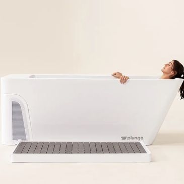Woman relaxing in a sleek, modern white bathtub with a wooden bath mat.