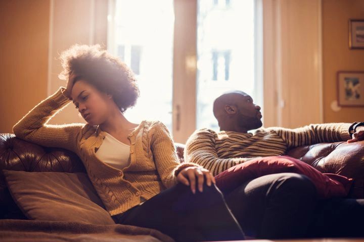 A couple sits apart on a couch looking upset and distant.