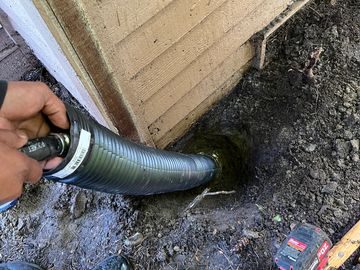 Inserting a black drainage pipe into a hole near a house's foundation.