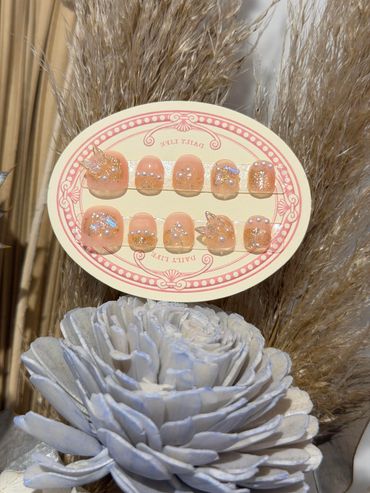 Elegant peach-colored press-on nails with pearl and glitter embellishments.