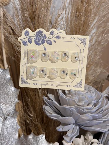 Elegant clear press-on nails with sparkling embellishments on a decorative card.
