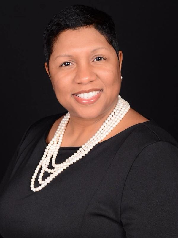 Smiling woman wearing a black top and layered pearl necklace against a black background.