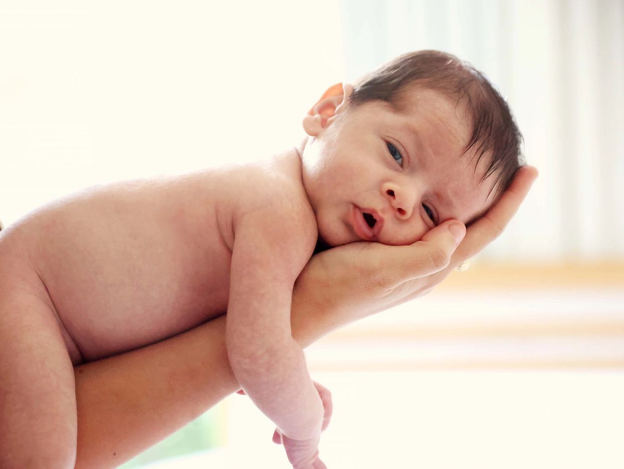 Newborn baby resting on an adult's hand with a curious expression.