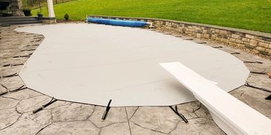 Covered swimming pool with diving board on a stone patio in a backyard.
