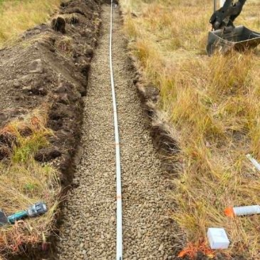 Trench filled with gravel and pipe installation in grassy field.