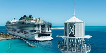 MSC World America docked near a lighthouse with two people on the lighthouse balcony.