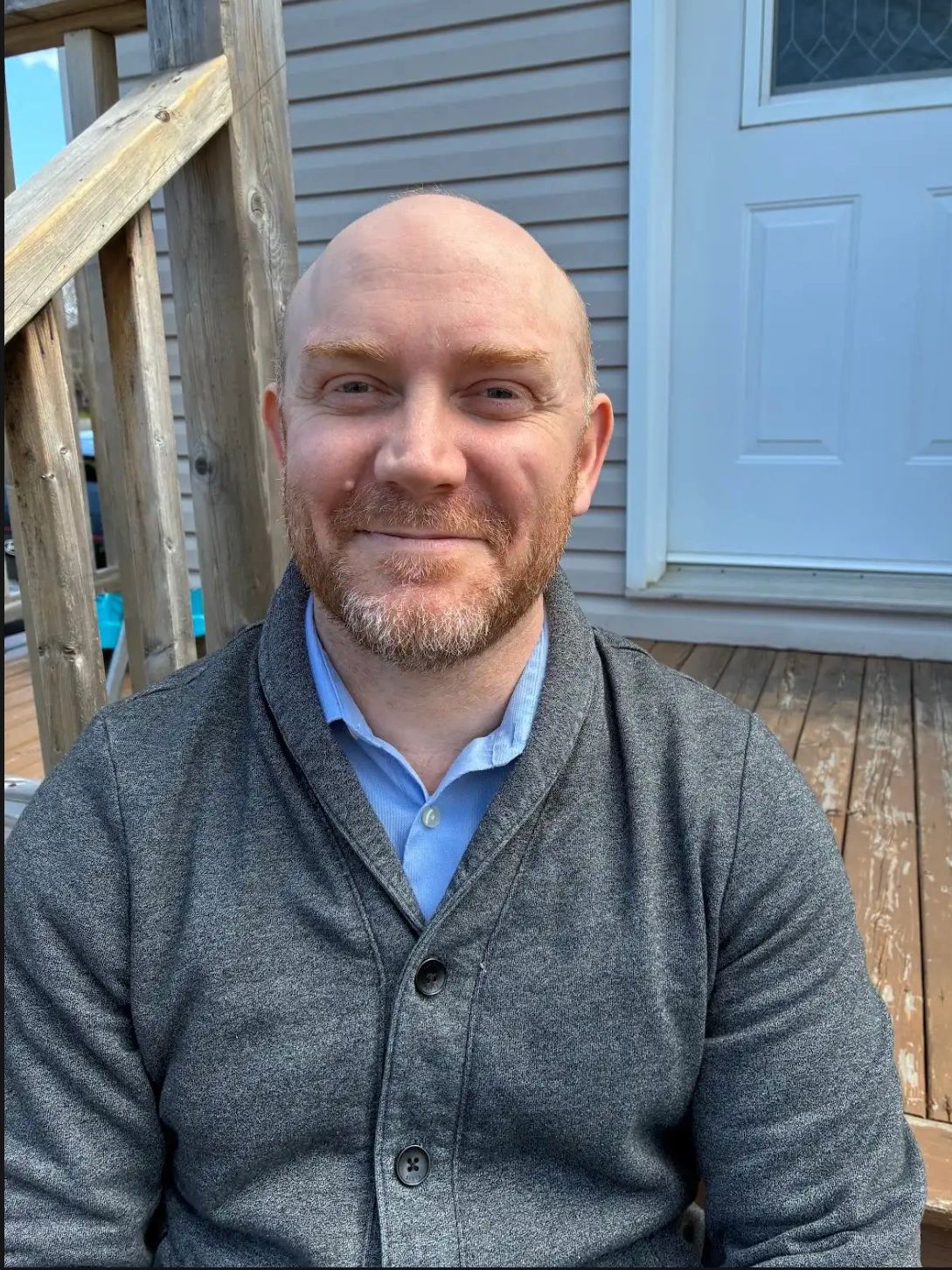 Smiling man with beard sitting on a wooden porch in casual attire.