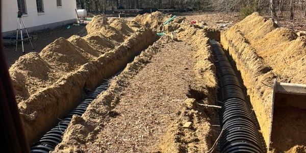 Trenches with large black drainage pipes beside a white house under a clear blue sky.