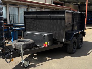 Black utility trailer with dual axles and spare tire parked outside a workshop.