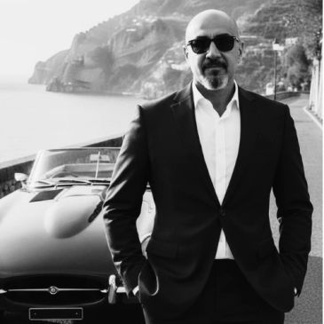 Man in sunglasses and suit standing by a vintage car with mountainous coastline in background.