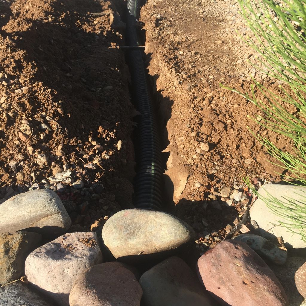 Black drainage pipe installed in a dirt trench surrounded by rocks and plants.