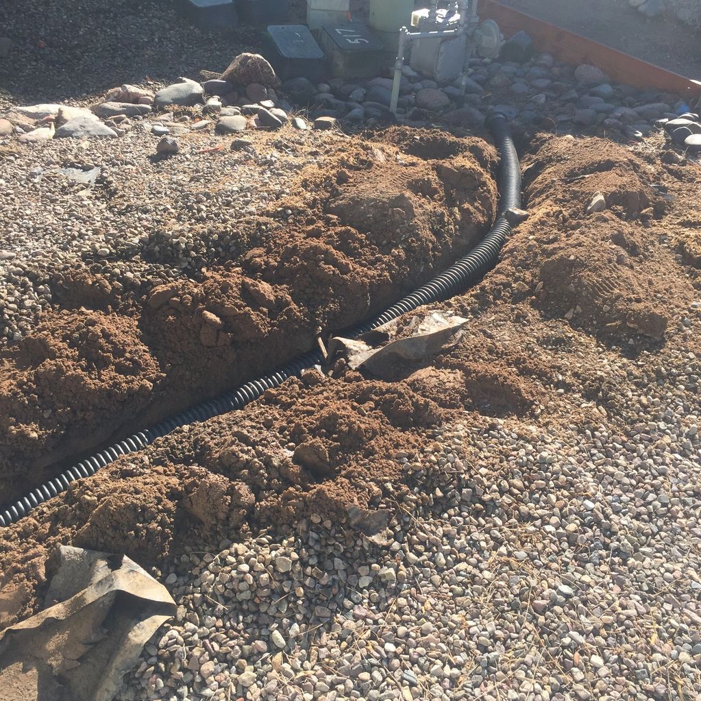 A black corrugated pipe partially buried in a trench with surrounding dirt and gravel.