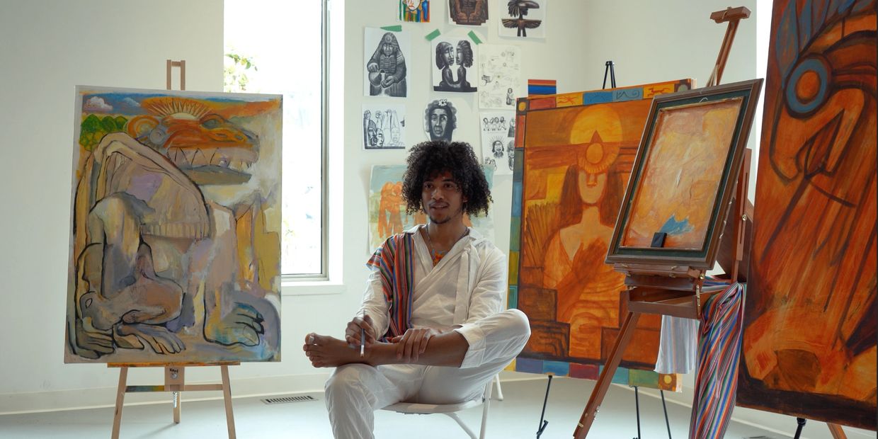 Artist sitting surrounded by colorful paintings and sketches.