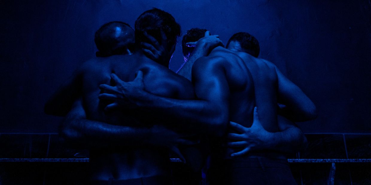 Group hug among four shirtless men under blue light.