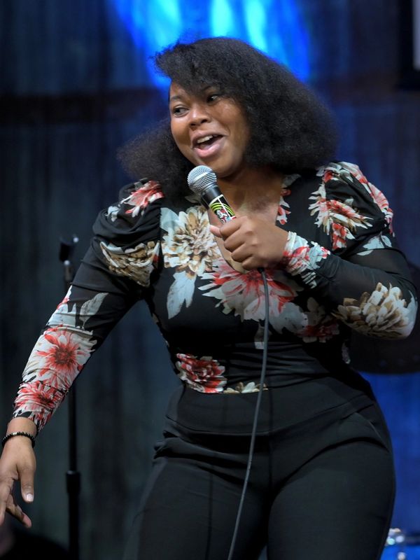 Woman in floral top performing stand-up comedy with a microphone.