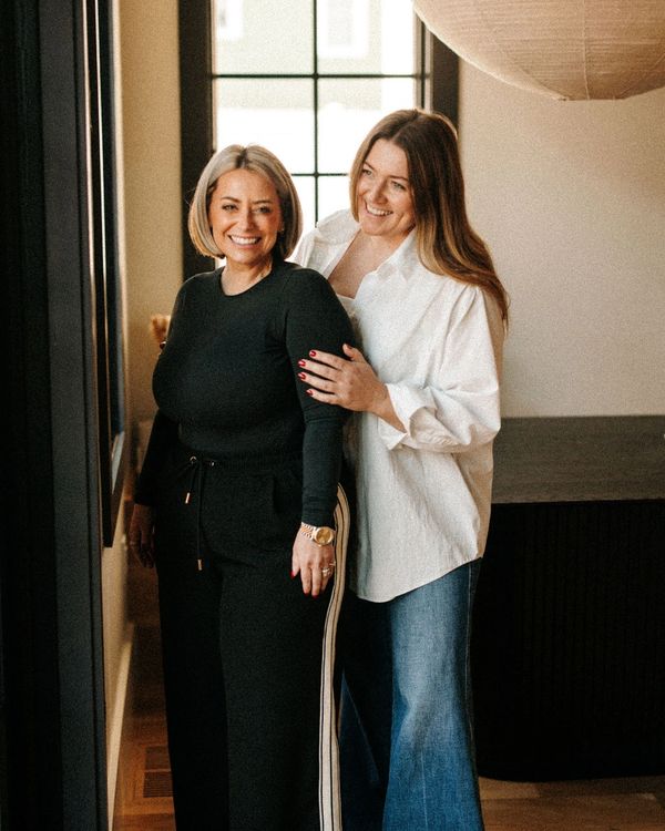 Two smiling women standing close together by a window in a cozy room.