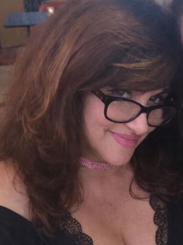Woman with glasses smiling, wearing a pink choker and black lace top.