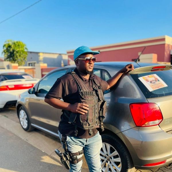 Man in tactical vest and cap leaning on a car, holding a drink.