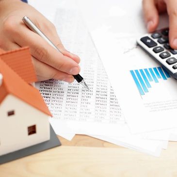 A hand holding a pen over documents with numbers and charts next to miniature house and calculator