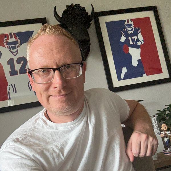 Man in glasses and white shirt takes selfie in room with sports art and buffalo head decor.